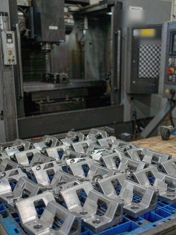 Production batch of identical machined aluminum brackets arranged in a blue crate beside a CNC machining center