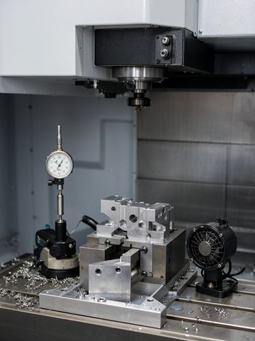 Machined aluminum part clamped in a precision vise on a CNC machine table with a dial indicator set up beside it