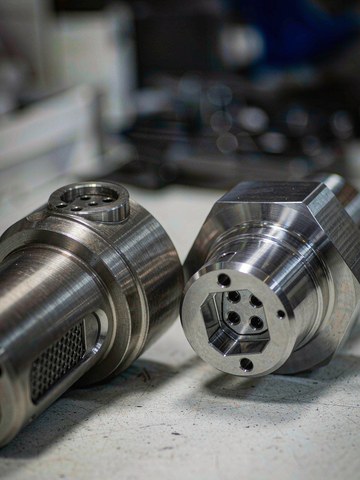 Two machined stainless steel fittings with different surface finishes side by side on a workbench