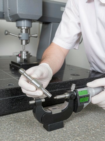 Quality inspector measuring a machined metal part with a digital micrometer beside a granite bridge CMM